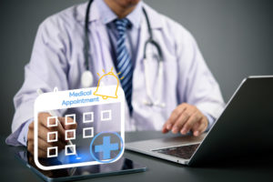 Agendamento online: principais benefícios para transformar sua clínica 1 Médico sentado em seu consultório usando o seu notebook e tablet para marcar consultas