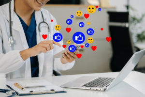 Médica sentado em frente a computador usando celular com ícones de redes sociais em volta