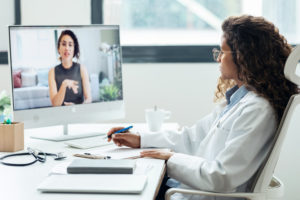 Como a Teleconsulta do iClinic ajuda a Dra. Verônica a fidelizar seus pacientes? 1 Médica realizando um atendimento à distância em seu consultório usando um computador