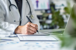 7 motivos para adotar uma agenda médica personalizável no consultório 1 Médico de jaleco branco escrevendo em papel e configurando agenda médica personalizável no computador