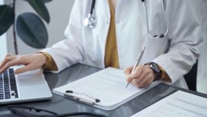 Médico sentado em frente a um computador faz anotações em um papel
