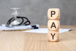 Salário de secretária de consultório médico: fatores para avaliar antes de defini-lo 1 Blocos de madeira empilhados formando a palavra “PAY” com estetoscópio e ampulheta ao fundo