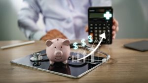 Implementando processos administrativos eficientes: do agendamento ao faturamento 1 Médico segurando uma calculadora logo atrás de um tablet com um porquinho, um estetoscópio e um gráfico ascendente
