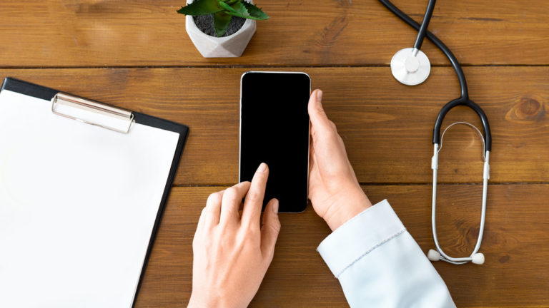 Médica segurando um celular em uma mesa com vários itens a sua volta
