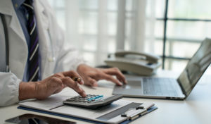 Precificação de procedimentos: como definir valores com base em custos e demanda 1 Médico de frente para um notebook fazendo um cálculo em uma calculadora