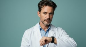 Médico de jaleco branco com um relógio no pulso com expressão de cansado